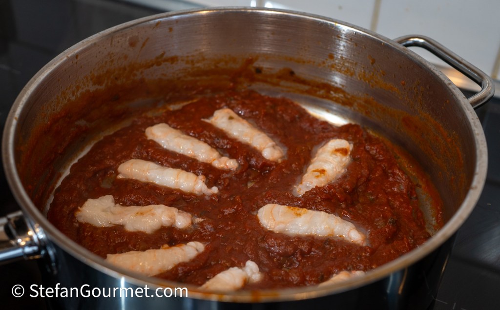 A metal pot containing a rich red sauce with several pieces of fish submerged in it, simmering on a stovetop.