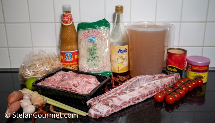 An assortment of ingredients for cooking, including rice noodles, ground meat, pork ribs, bean sprouts, shallots, garlic, tomatoes, and various sauces on a kitchen counter.