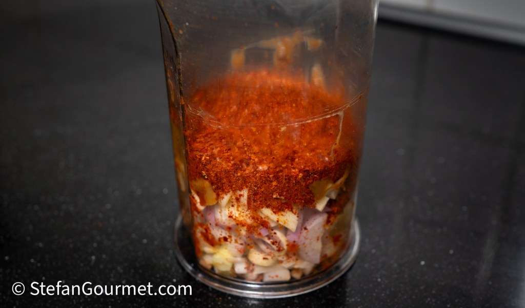 A clear container filled with chopped garlic and onion, topped with a layer of red chili powder, placed on a black countertop.