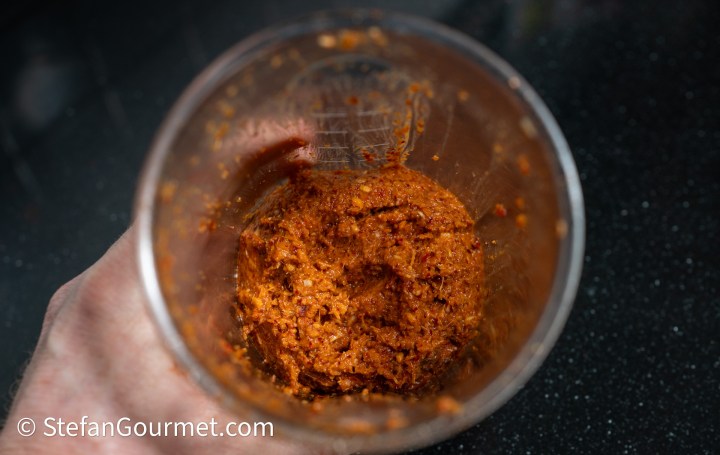 A hand holding a glass jar containing a thick, reddish-brown paste or sauce, with a textured surface, on a dark countertop.
