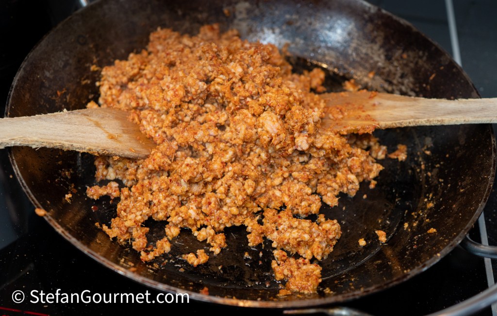 A close-up of a frying pan filled with cooked, crumbled sausage mixture being stirred with a wooden spoon.