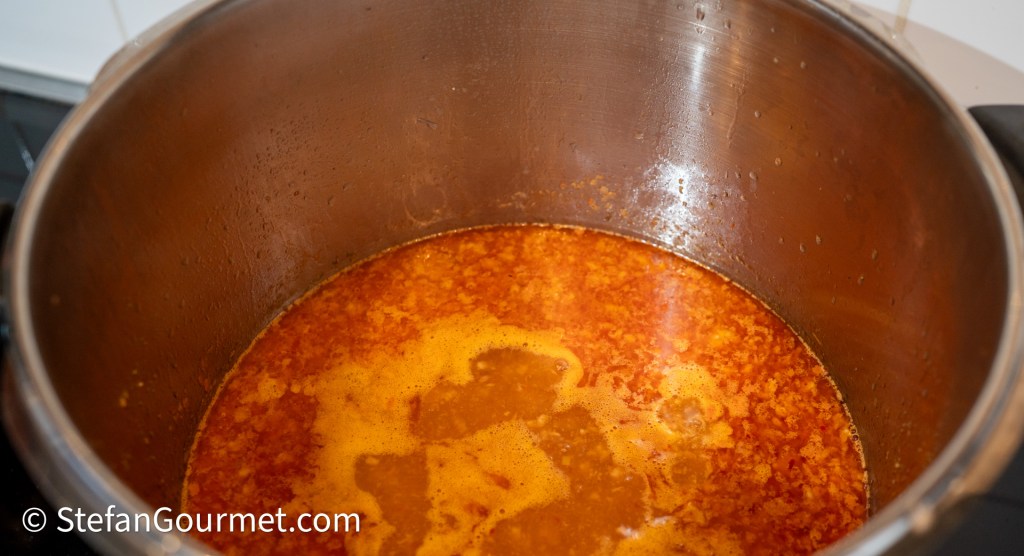A close-up view of a pot containing simmering orange broth, with bubbles on the surface and steam rising, set on a stovetop.