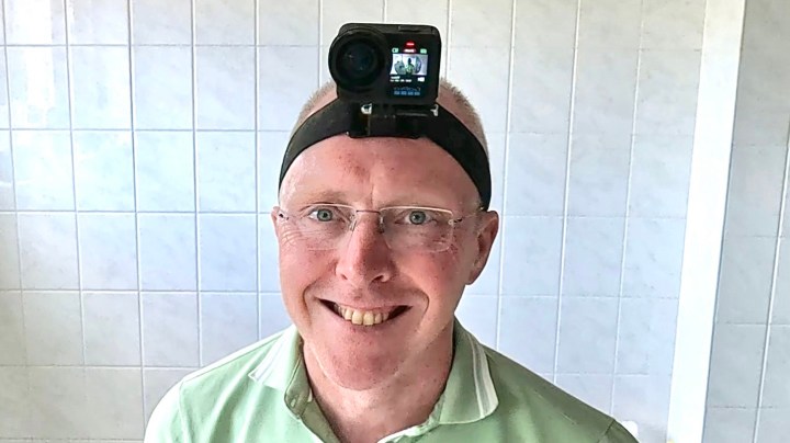 A smiling man wearing a green shirt and glasses, with a camera mounted on a headband on his forehead, standing in a tiled room.