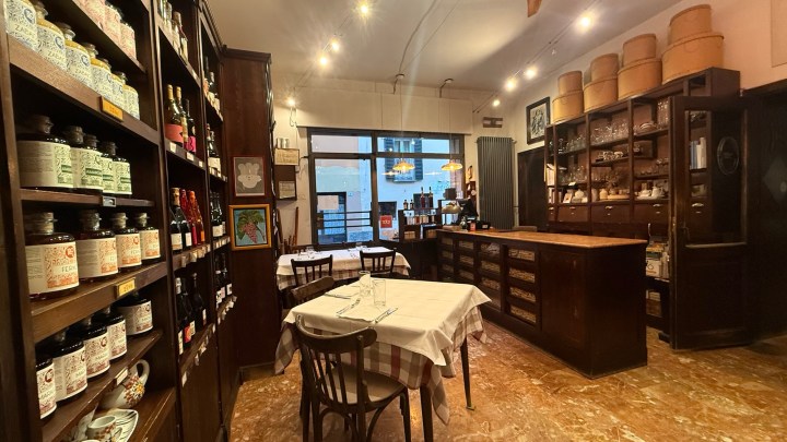 Interior of a quaint restaurant featuring wooden shelves filled with bottles and jars, a table set for dining, and a bar area in the background.