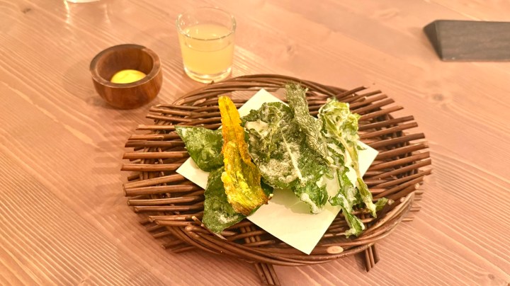A plate of crispy fried vegetables including leafy greens and zucchini blossoms, served on a woven basket with a small bowl of dipping sauce and a glass of light-colored beverage.