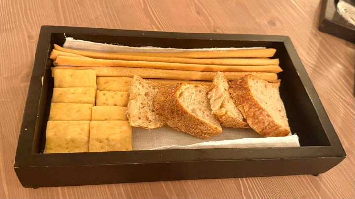 A wooden tray containing various types of bread, including sliced brown bread and rectangular crackers, alongside long breadsticks.