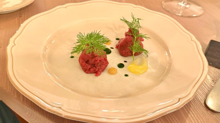 A gourmet dish featuring two spherical portions of red meat topped with fresh dill on a decorative plate, accompanied by colorful sauces including yellow and green dots.