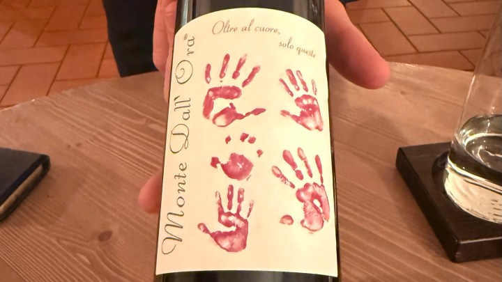 A bottle of wine labeled 'Monte Dall'Ora' featuring red handprint designs on the label, held by a person against a wooden surface.