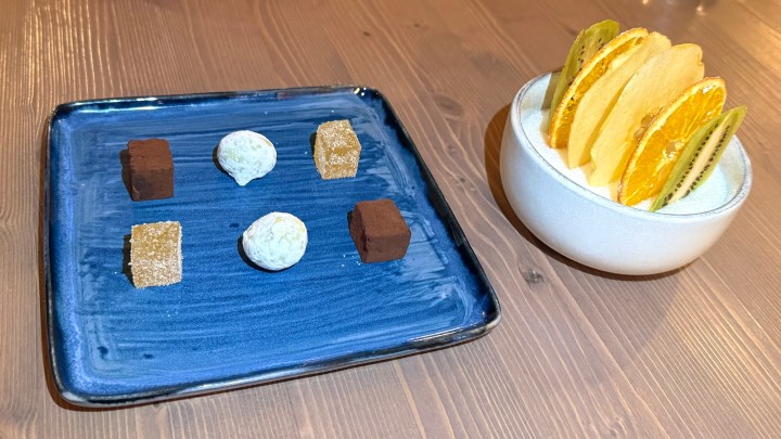 A blue plate featuring an assortment of confections, including chocolate squares, round candies, and sugar cubes, arranged in two groups. Adjacent to the plate, a white bowl contains sliced fruits such as oranges and kiwis.