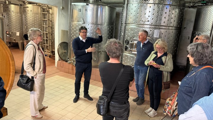 A group of people gathered in a winery, listening to a guide speaking near stainless steel fermentation tanks.