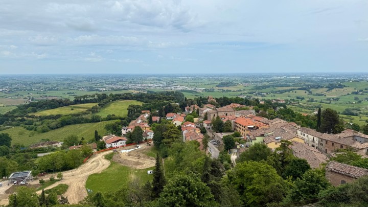 A panoramic view of a lush green landscape featuring a quaint village with terracotta-roofed houses nestled among trees, rolling hills, and expansive fields under a cloudy sky.