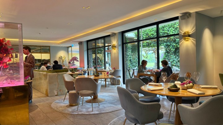 A modern restaurant interior with elegant decor, featuring round tables and stylish chairs. Guests are dining and socializing, while large windows let in natural light and show greenery outside.