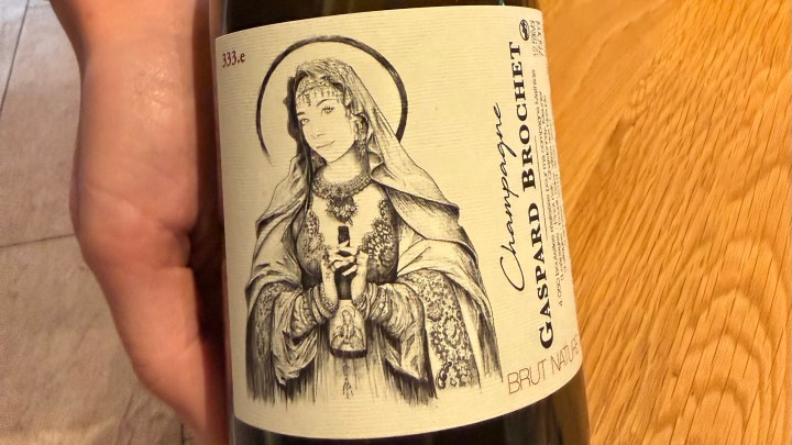 A hand holding a champagne bottle with an artistic label featuring a woman in traditional attire, labeled 'Champagne Gaspard Brochet Brut Nature'.
