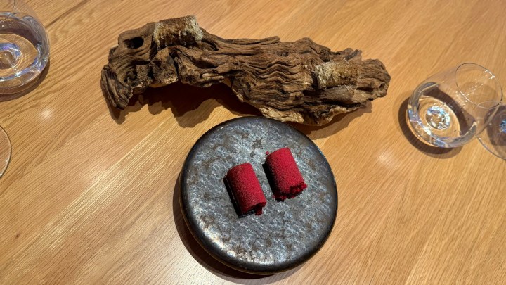 A dish featuring two red food items placed on a round dark stone plate, accompanied by a piece of rustic wood and glassware on a wooden table.