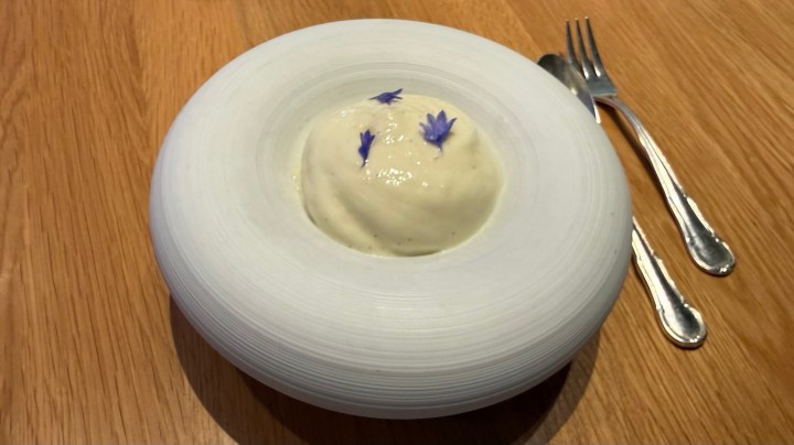 A round white dish containing a creamy dessert topped with small blue edible flowers, placed on a wooden table beside silver cutlery.