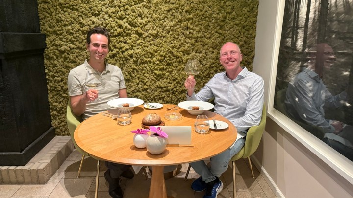Two men sitting at a round wooden table in a restaurant, enjoying a meal. One man is holding a glass, while the other is gesturing with a utensil. They both have plates in front of them, and there are decorative elements on the table.