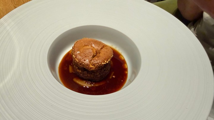 A round dessert soufflé served in a white plate, sitting in a small pool of dark sauce.
