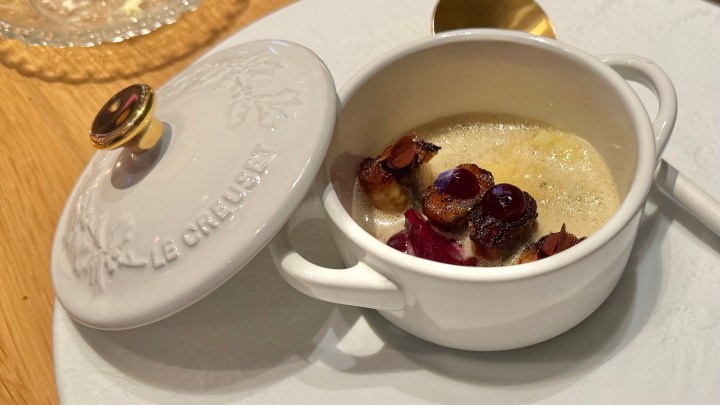 A white ceramic dish with a lid, containing a gourmet dish featuring small pieces of food garnished with colorful elements, placed on a wooden table.