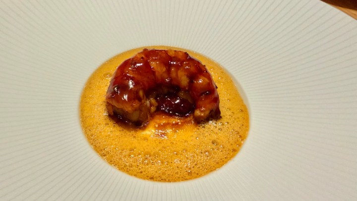 A gourmet dish featuring a caramelized seafood element presented in a shallow bowl with a frothy sauce surrounding it.