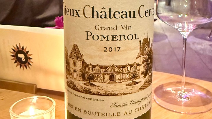 A bottle of Vieux Château Certan Grand Vin Pomerol 2017 with an illustration of the chateau on the label, placed on a wooden table next to a candle and wine glass.