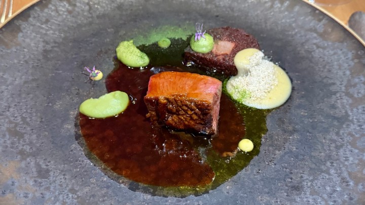 A gourmet dish featuring two pieces of seared meat on a dark plate, surrounded by various green and yellow sauces and garnishes.