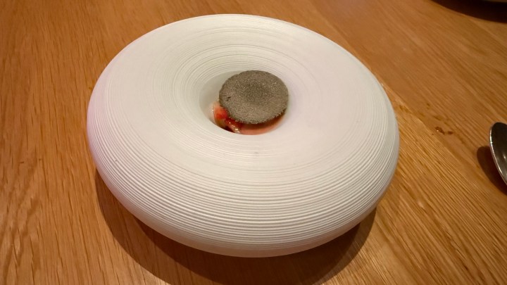 An artistic white plate with a concentric circular design, featuring a small grey circular element in the center and hints of red visible underneath.