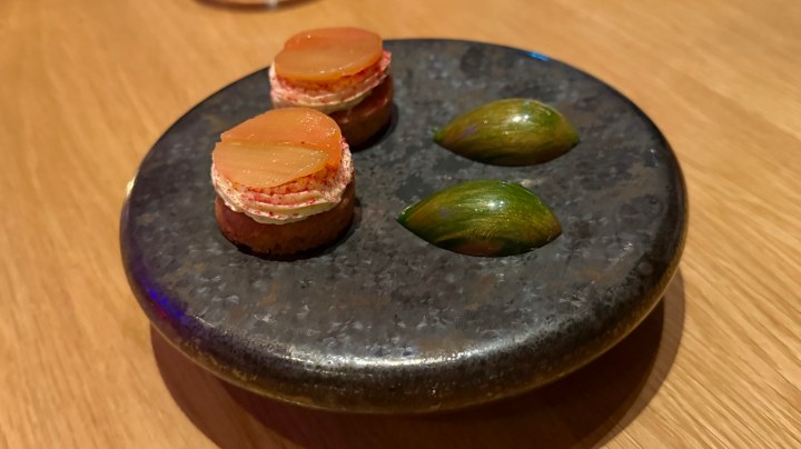 A dark stone plate featuring two small layered pastry bites topped with vegetables, alongside two green shellfish.
