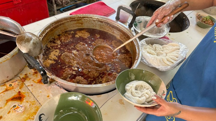 A chef ladling rich broth from a large pot into a bowl containing noodles, with various ingredients prepared in the background.