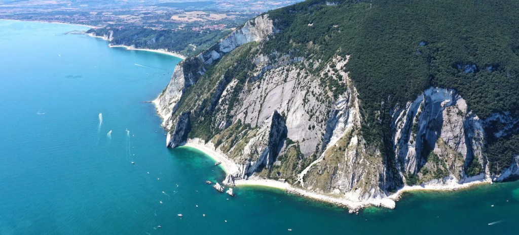 Aerial view of a coastline featuring steep cliffs, dense greenery, and a vibrant turquoise sea with small boats scattered in the water.