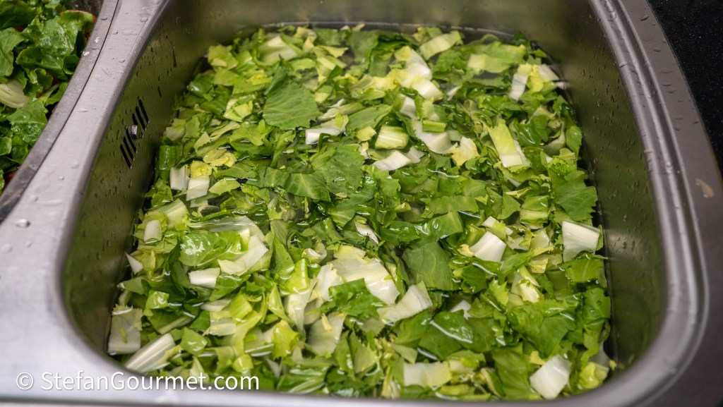 A stainless steel sink filled with chopped leafy greens, including lettuce and other salad vegetables, submerged in water.