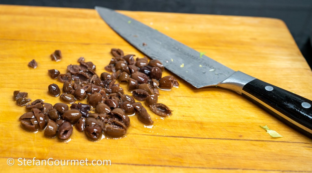 Chopped black olives on a wooden cutting board next to a chef's knife.