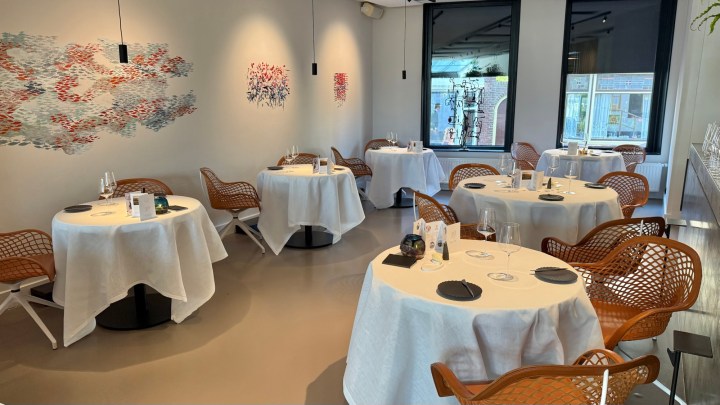 Interior of a modern restaurant featuring tables with white tablecloths and stylish wicker chairs, decorated with colorful wall art.