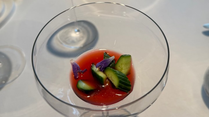 A glass dish containing a red sauce with sliced cucumbers and purple edible flowers on a white tablecloth.