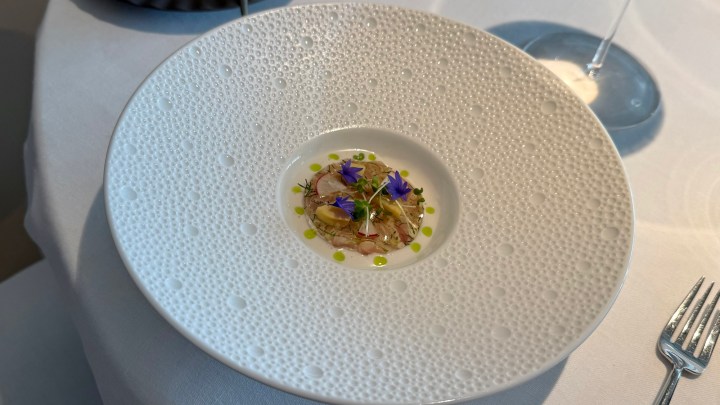 A gourmet dish presented on a textured white plate, featuring thinly sliced seafood garnished with edible flowers and green drizzles.