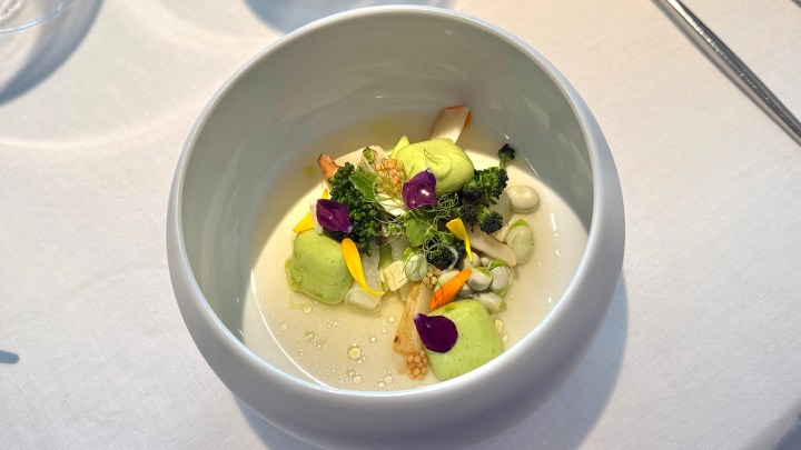A gourmet dish served in a white bowl, featuring green-colored foams, assorted vegetables, edible flowers, and a light broth.