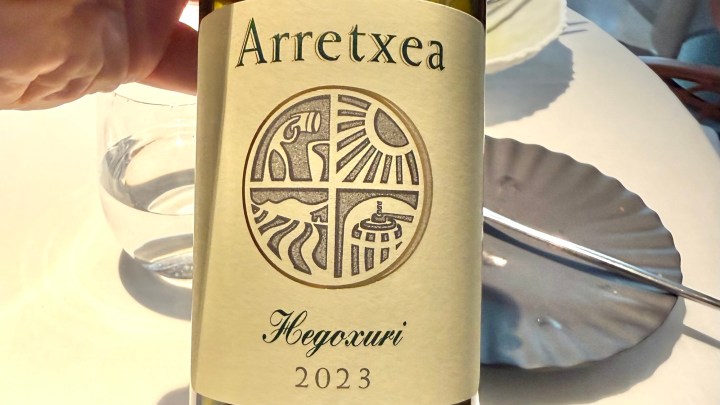 A close-up of a wine bottle label featuring the name 'Arretxea' and 'Hegoxuri 2023', with a decorative emblem divided into four sections, set against a light background with a glass of water nearby.