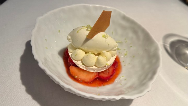 A delicate dessert featuring a scoop of ice cream atop a layer of strawberries, garnished with dollops of cream and a chocolate shard, presented in a white dish.