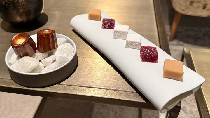 A plate with colorful bite-sized confections, including squares of different flavors, displayed alongside a small bowl of white stones and two canelé pastries.