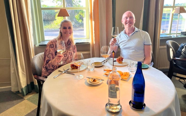 A man and a woman sit at a dining table, raising glasses in a toast. They are enjoying a meal with plates and drinks on the table, featuring a bright and airy atmosphere with windows in the background.