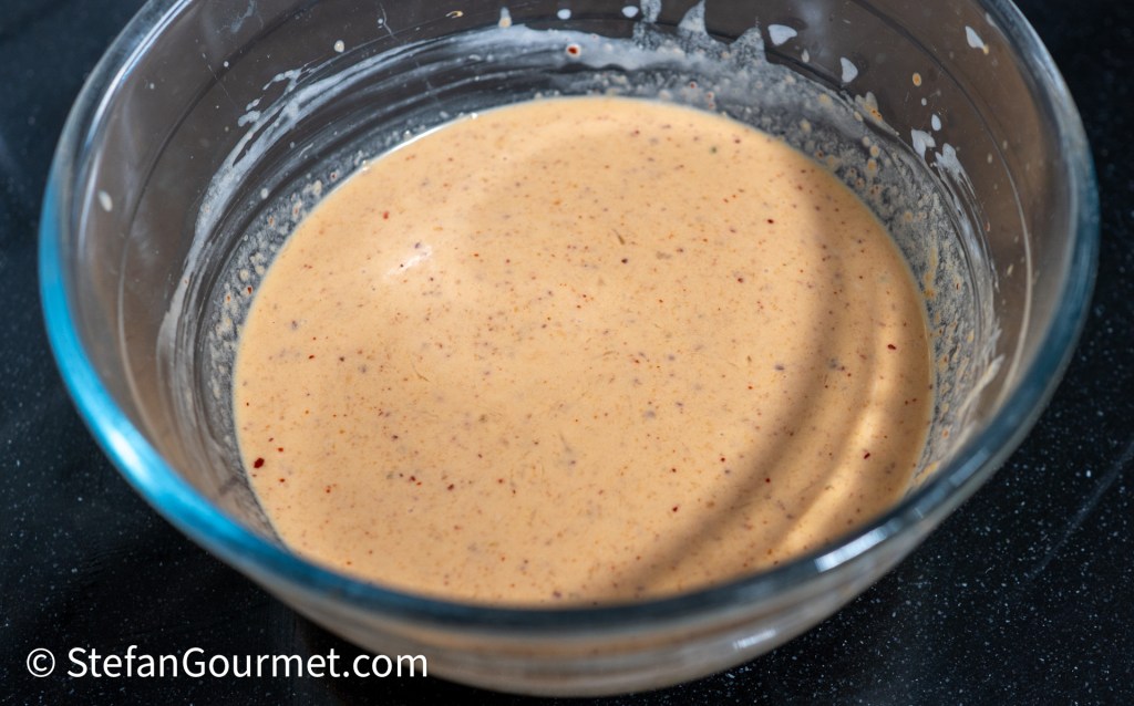 A clear glass bowl containing a creamy, light brown sauce or mixture with a smooth texture and small flecks of spice.