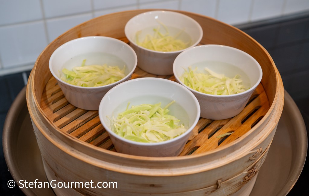 Four bowls with shredded cabbage in water, placed inside a bamboo steamer.