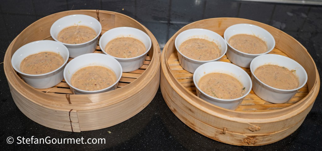 Two bamboo steamers containing small bowls filled with a light brown sauce or dip, placed on a black countertop.