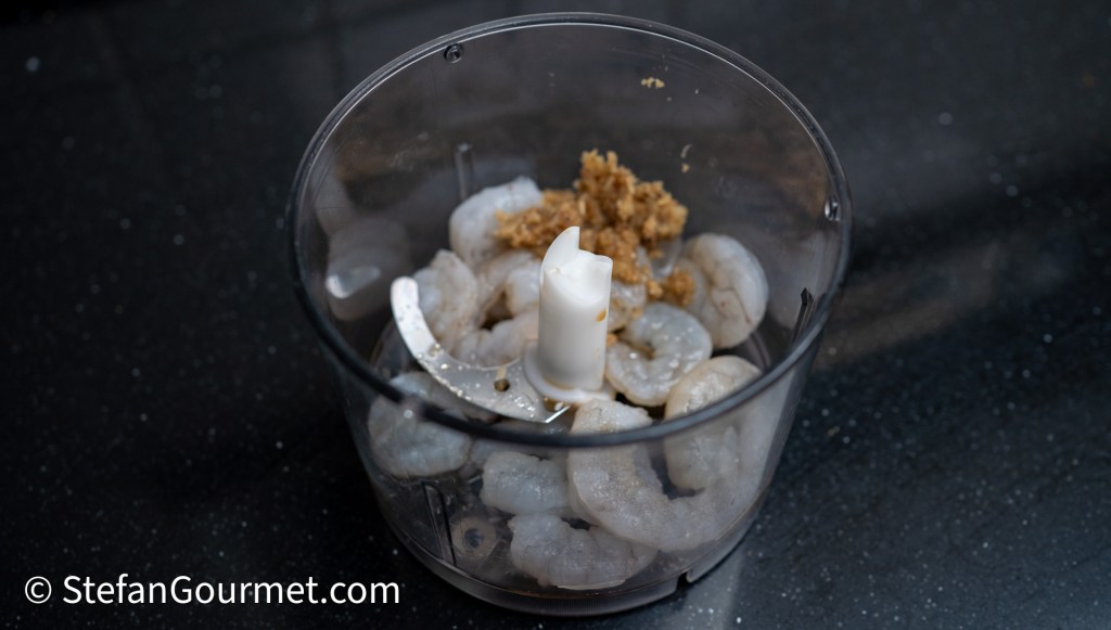 A food processor bowl containing raw shrimp and a small amount of brown sugar, ready for blending.