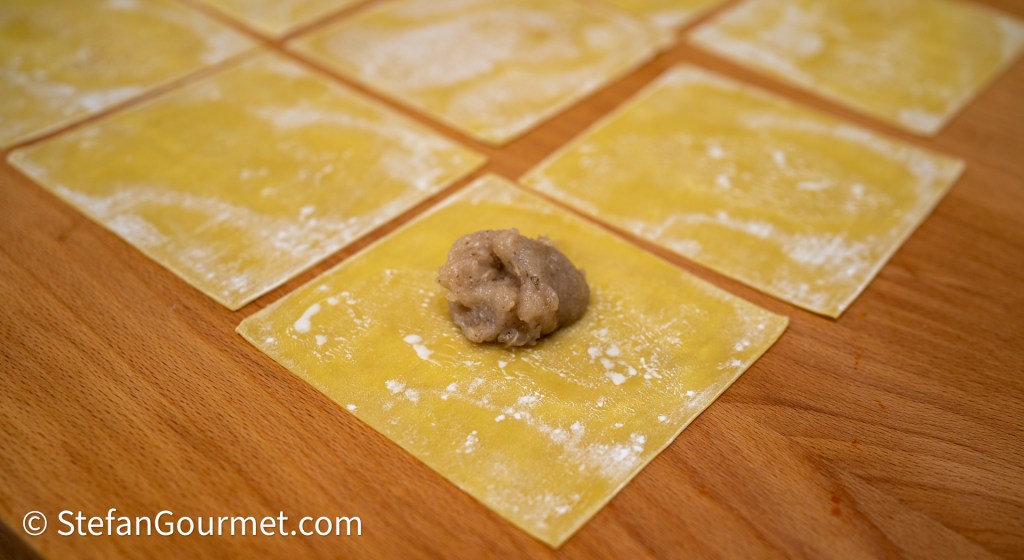 A wooden surface with several sheets of yellow pasta dough, one sheet featuring a small mound of filling placed in the center.