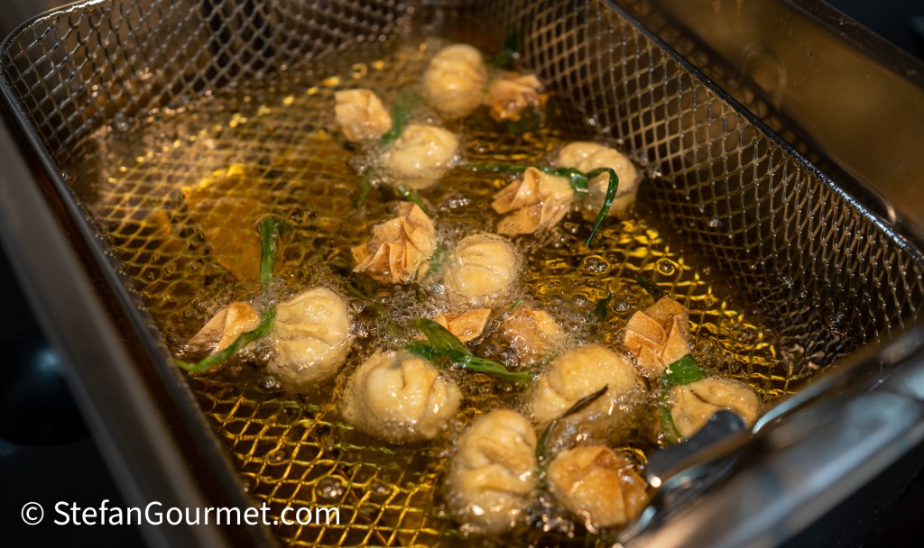 A deep fryer filled with bubbling oil and frying dumplings, garnished with green herbs.