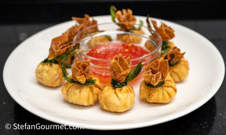 An elegant platter of golden, fried dumplings arranged in a circular pattern, each adorned with crispy decorations and green leaves. In the center, there is a small bowl of sweet chili sauce for dipping.