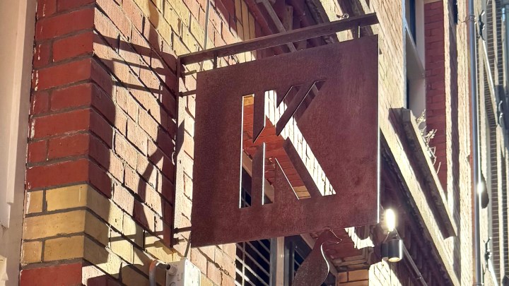 A metal sign displaying the letter 'K' hanging on a brick wall, with sunlight casting shadows.