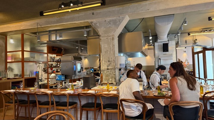 Interior view of a modern restaurant featuring an open kitchen, wooden bar seating, and diners enjoying their meals.