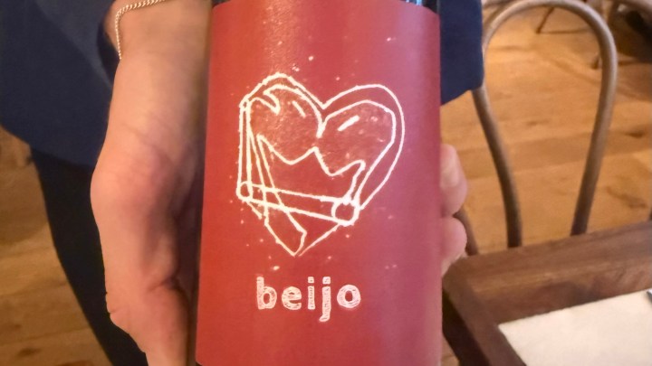 A person holding a wine bottle with a red label featuring a heart design and the word 'beijo' printed on it.