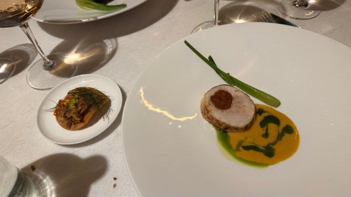 A plated gourmet dish featuring a round slice of meat topped with a condiment, accompanied by a decorative sauce swirl and a green garnish on a white plate. A small appetizer of a crispy base topped with herbs is positioned beside it.